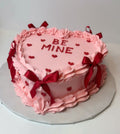 Heart-shaped pink cake with red ribbons and 'BE MINE' text on a white background