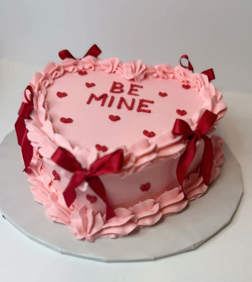 Heart-shaped pink cake with red ribbons and 'BE MINE' text on a white background