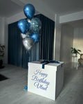 White gift box with 'Happy Birthday Vlad' text, blue and silver balloons in a room.