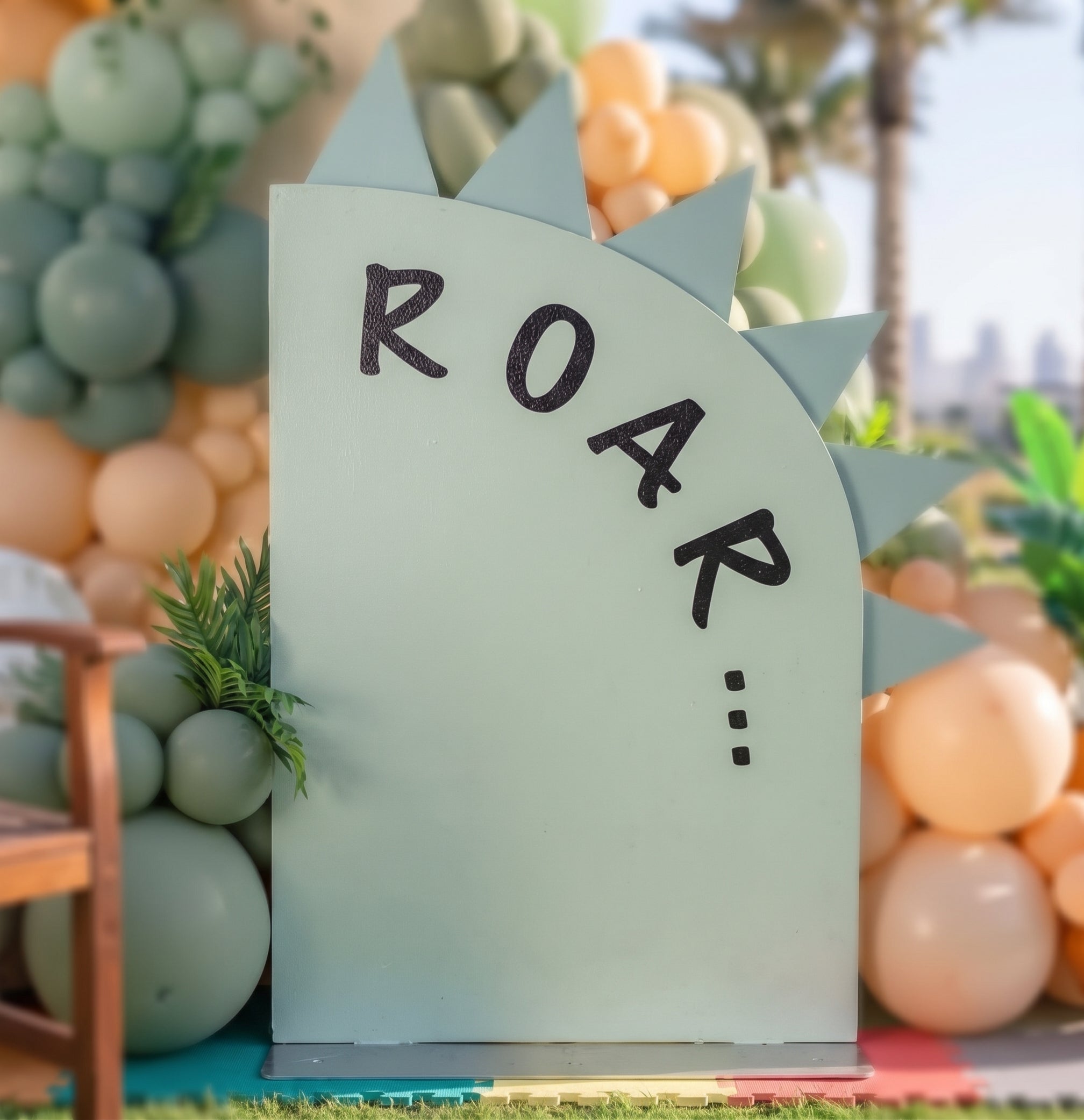 Dinosaur themed 1st birthday party backdrop setup in Dubai featuring arched wall, custom name text, sage green and orange balloon garland, and cute dinosaur cutouts.   