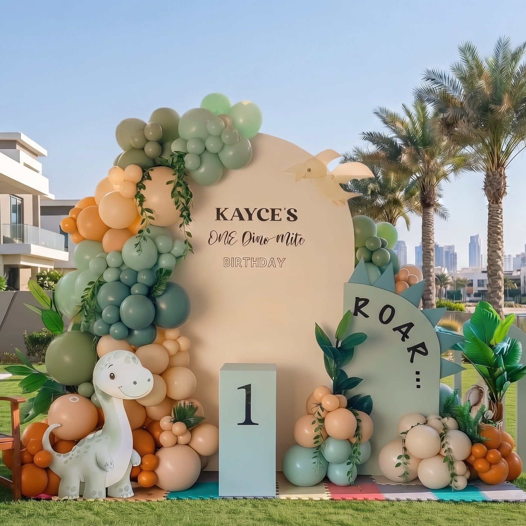 Dinosaur themed 1st birthday party backdrop setup in Dubai featuring arched wall, custom name text, sage green and orange balloon garland, and cute dinosaur cutouts.   