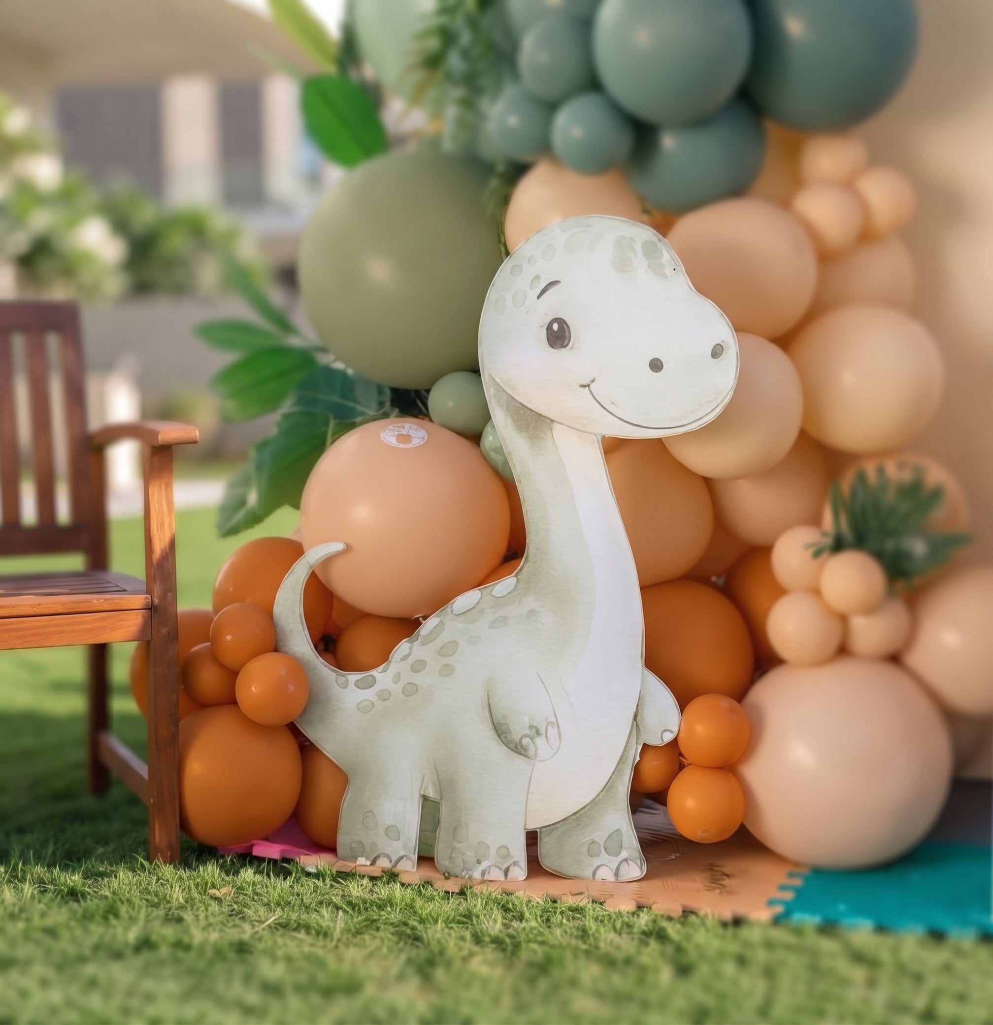 A white and light green dinosaur party decoration stands in front of a balloon garland backdrop with peach, orange, and sage green balloons, set on artificial grass with a wooden chair visible at the side.