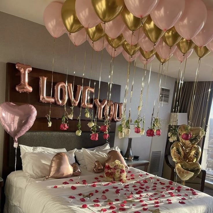 Romantic hotel bedroom decoration in Dubai with floating pink and gold helium balloons, hanging roses, rose gold foil letters, and rose petals on the bed.