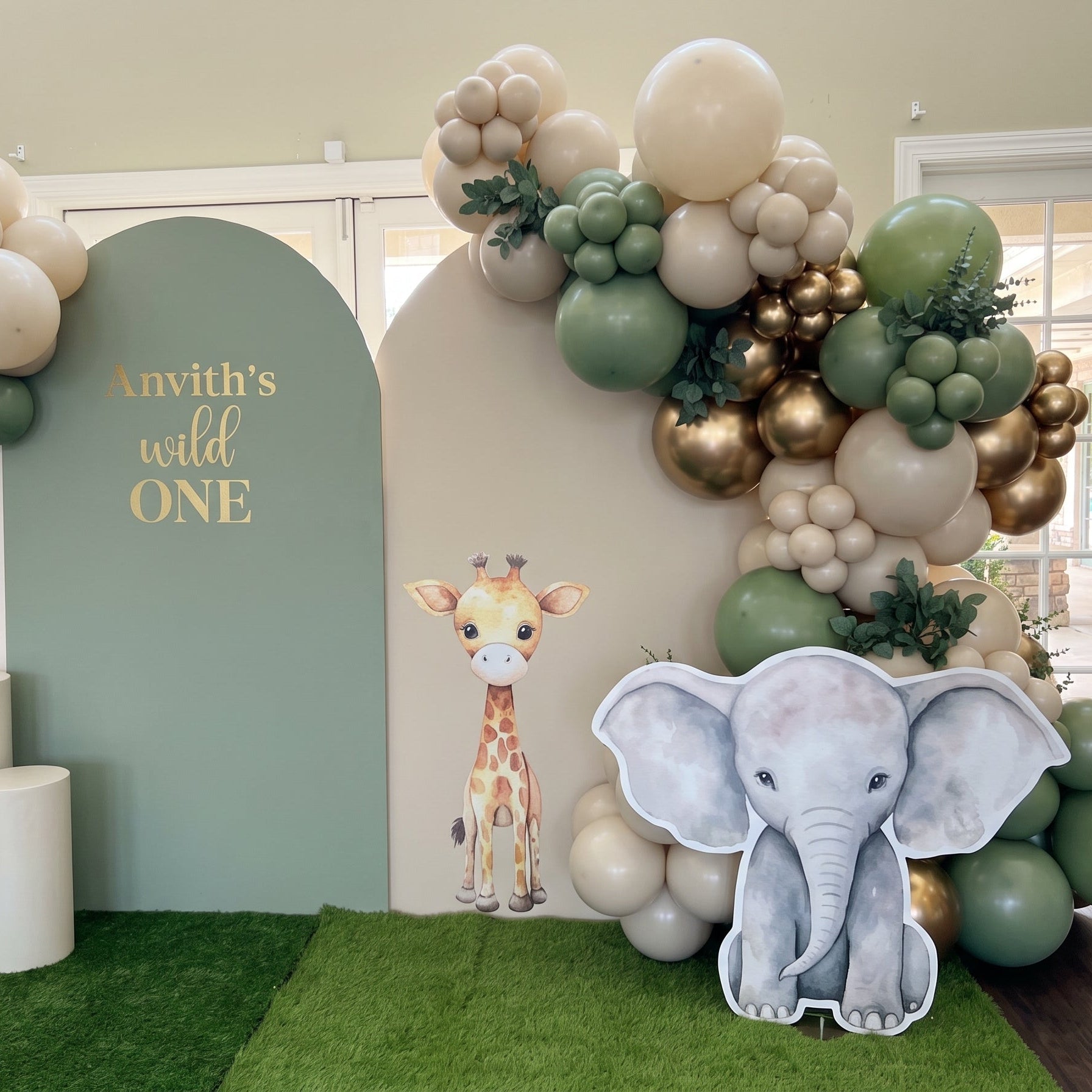 Wild One jungle safari 1st birthday stage decoration in Dubai featuring sage green arches, chrome gold balloon garland, and cute safari animal cutouts.