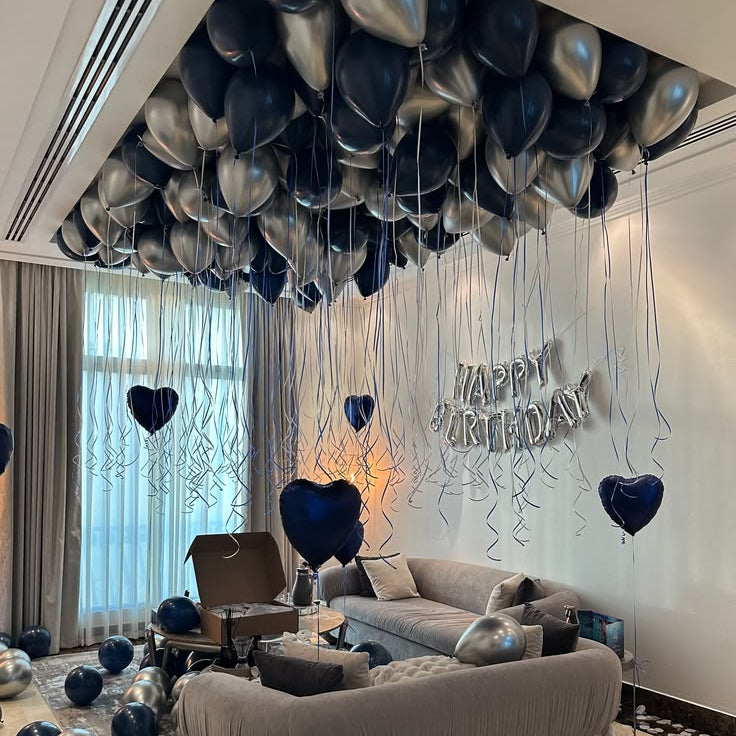 A styled birthday party setup in a living room with a large cluster of dark blue, navy, and metallic silver balloons hanging from the ceiling, with additional balloons scattered on the floor. A gray tufted sofa is positioned in the foreground, and a silver 'HAPPY BIRTHDAY' balloon sign is mounted on the wall.