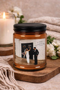 Candle jar with a printed label featuring a couple, placed on a wooden surface with flowers and another candle in the background.