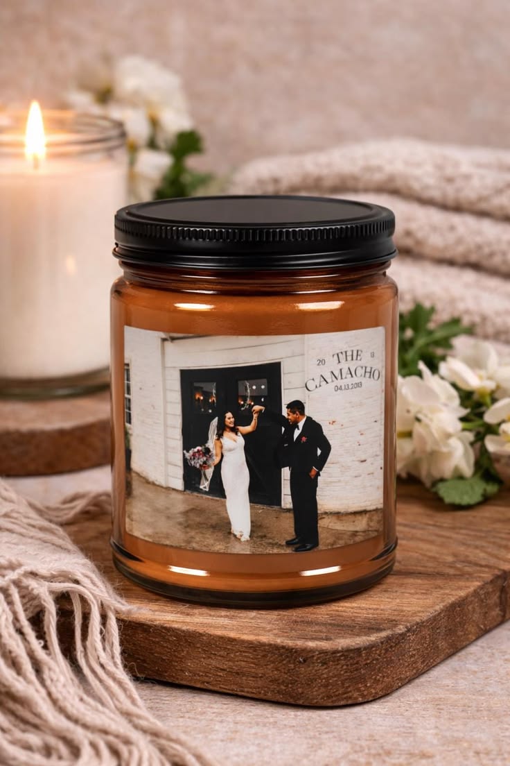 Candle jar with a printed label featuring a couple, placed on a wooden surface with flowers and another candle in the background.
