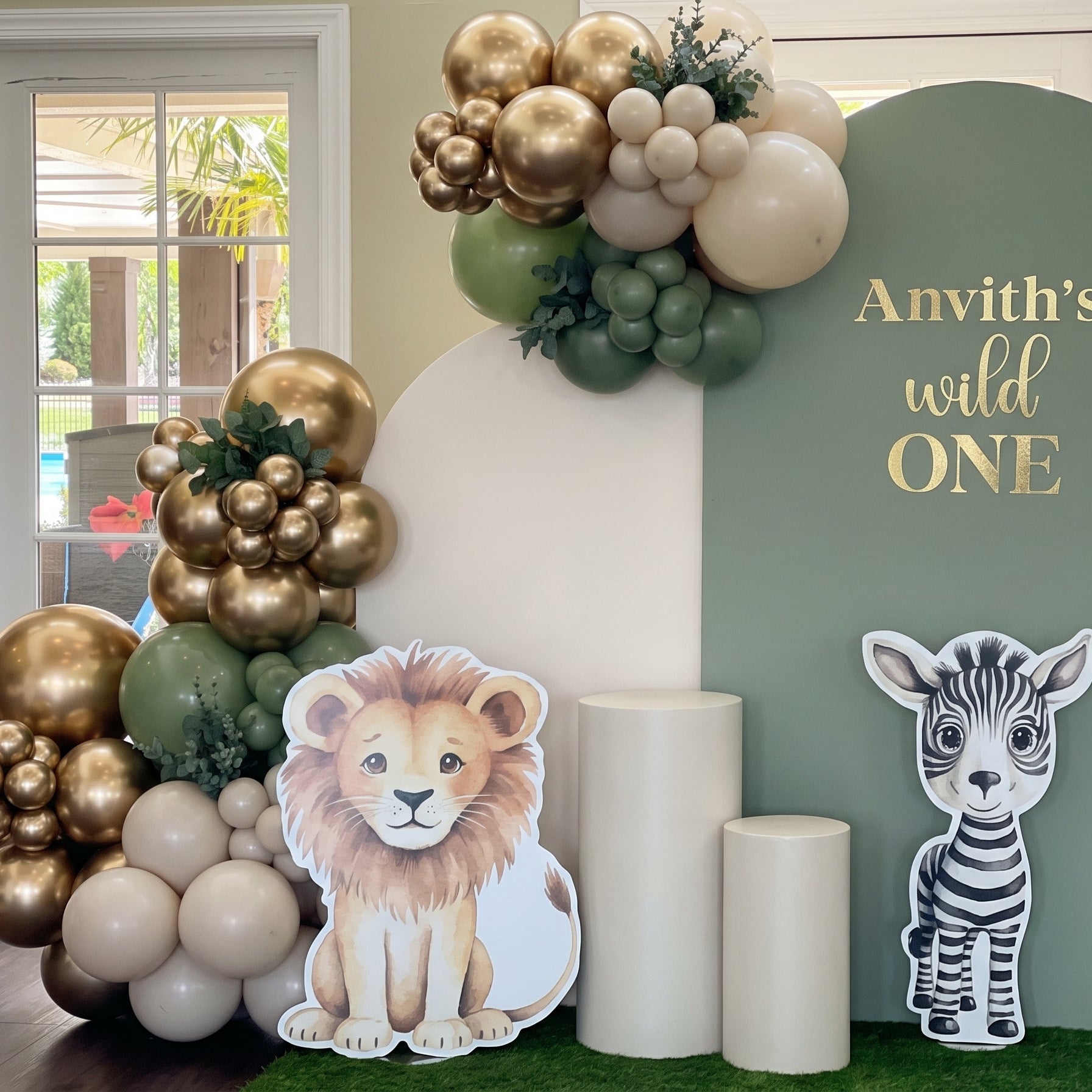 Wild One jungle safari 1st birthday stage decoration in Dubai featuring sage green arches, chrome gold balloon garland, and cute safari animal cutouts.