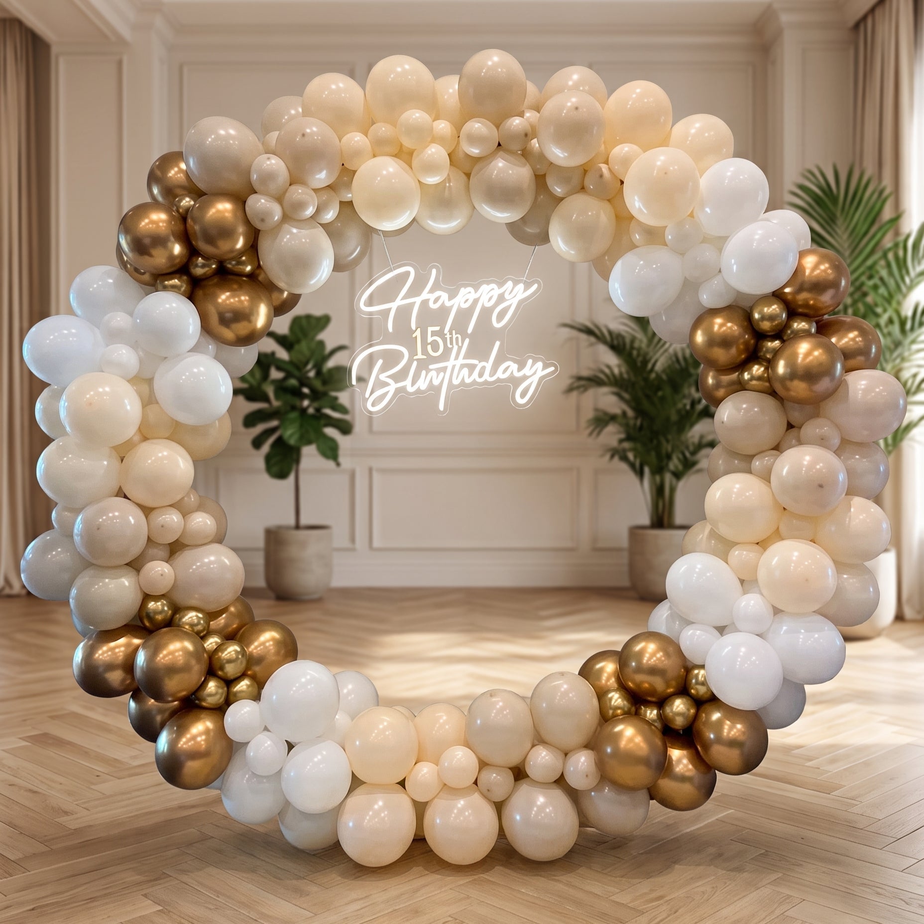 A large circular balloon garland decoration in champagne, white, and metallic gold, arranged in an arch on a wooden floor in a decorated room. The center features a glowing white script sign that reads 'Happy 15th Birthday'.