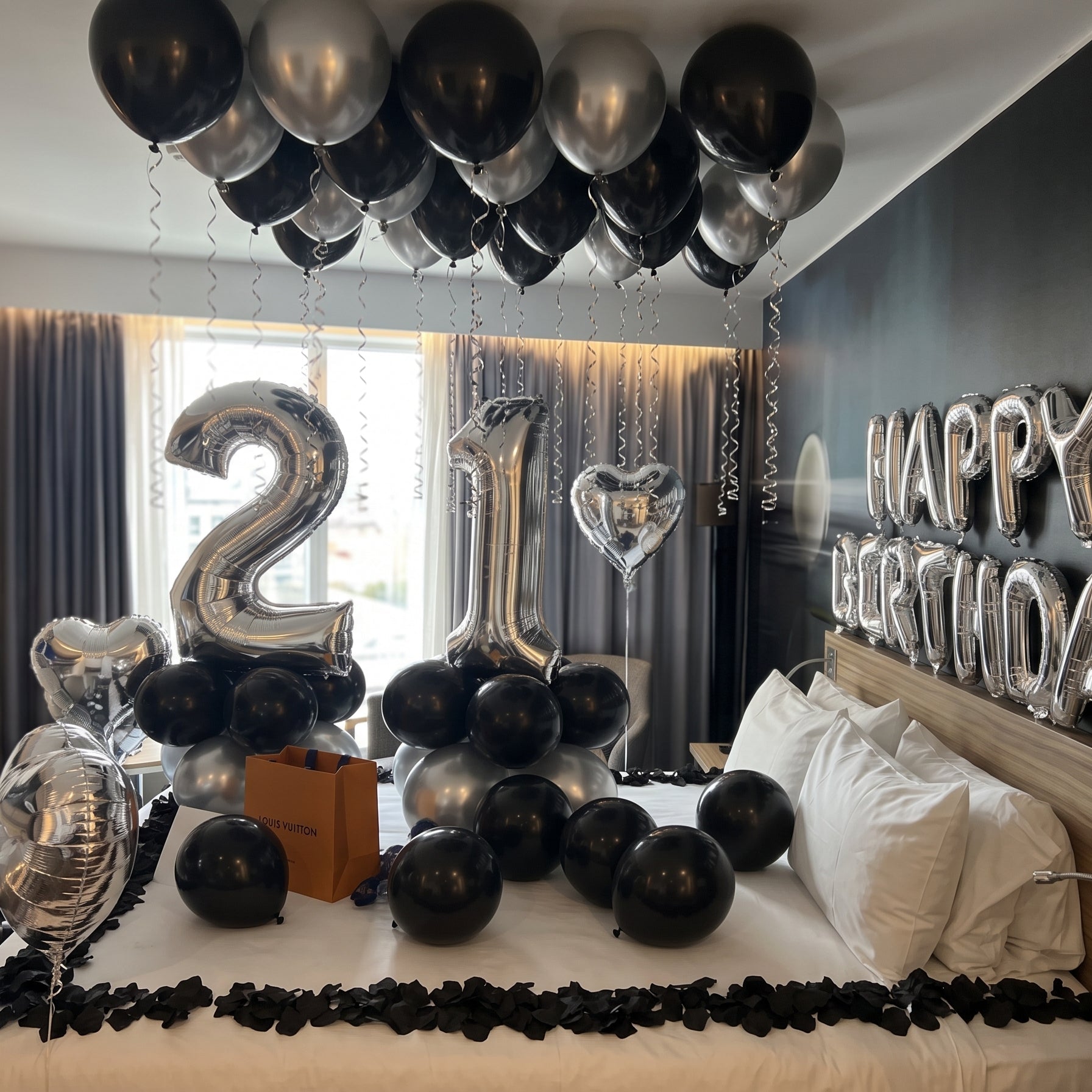 A bedroom decorated for a 21st birthday celebration, featuring a bed covered with black and silver balloons, a large metallic '21' balloon, a heart-shaped balloon, a gift box, and a 'HAPPY BIRTHDAY' balloon arrangement on the dark gray wall.
