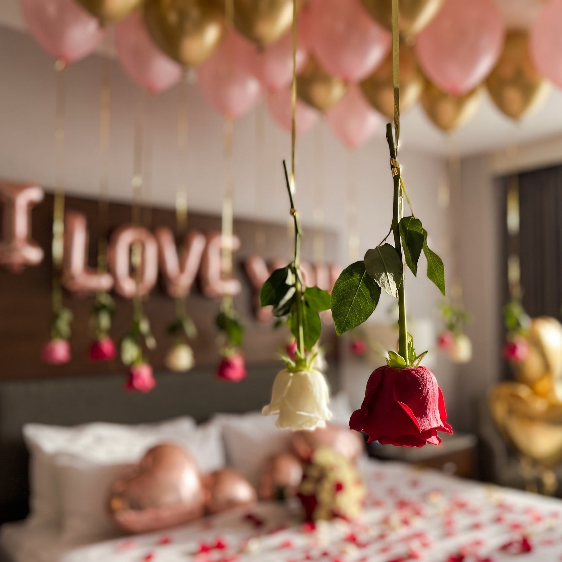 Romantic hotel bedroom decoration in Dubai with floating pink and gold helium balloons, hanging roses, rose gold foil letters, and rose petals on the bed.